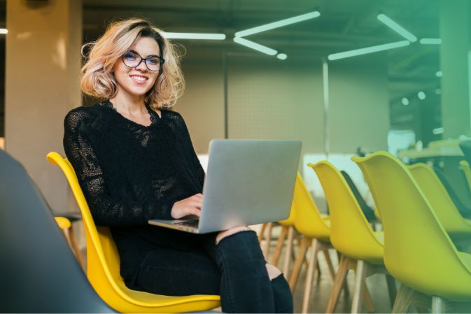 Blonde woman sitting on yellow chair and using her laptop in a big room