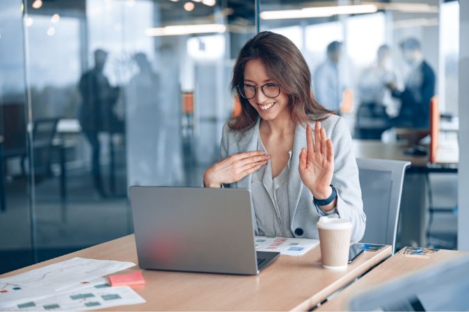 Frau, die in einem Büro vor einem Laptop sitzt und die Hand hebt, um anzuzeigen, dass sie in der Online-Sitzung etwas sagen möchte