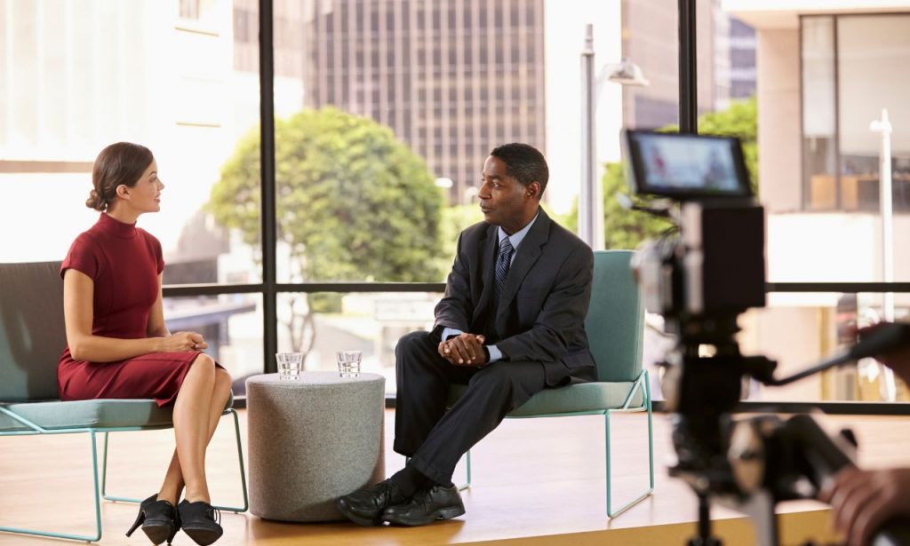 una mujer vestida de rojo entrevista a un hombre vestido de negro durante una retransmisión en directo con subtítulos en tiempo real