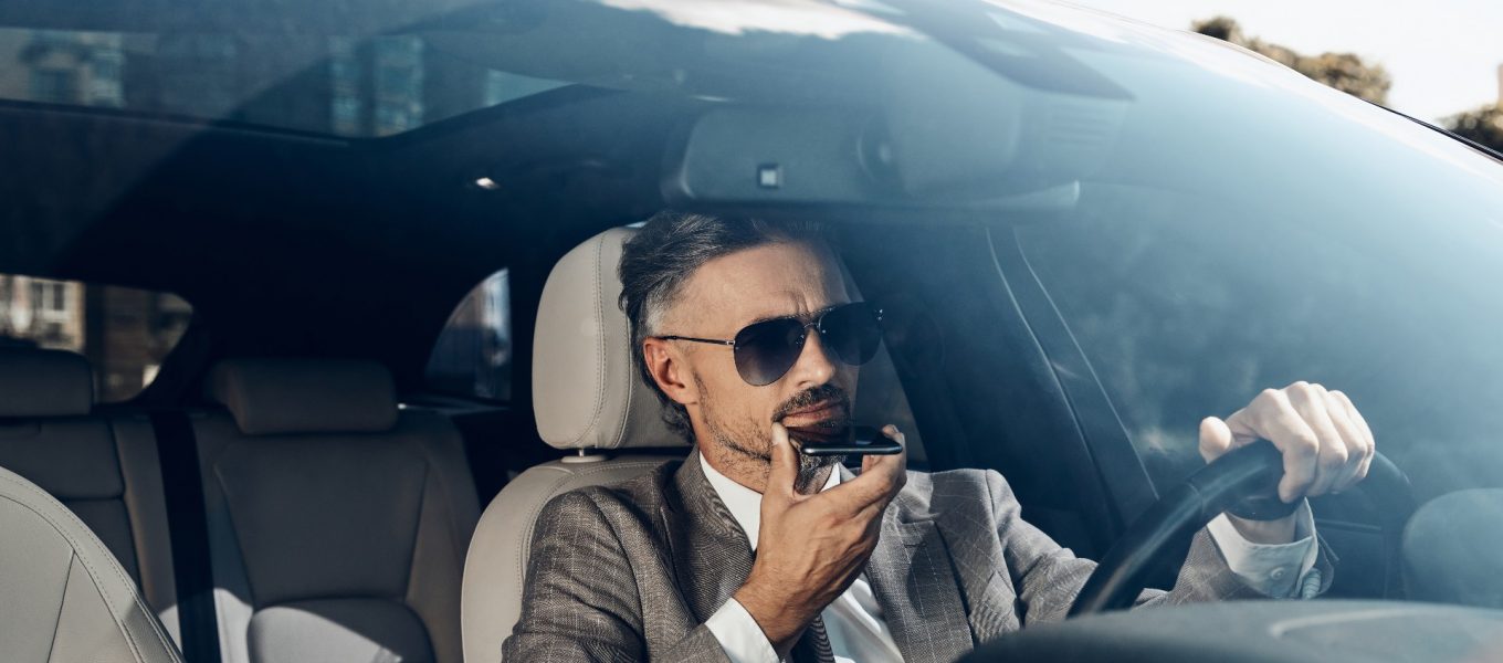 Geschäftsmann im grauen Anzug, der in seinem Auto sitzend eine App für die Umwandlung von Sprache in Text verwendet